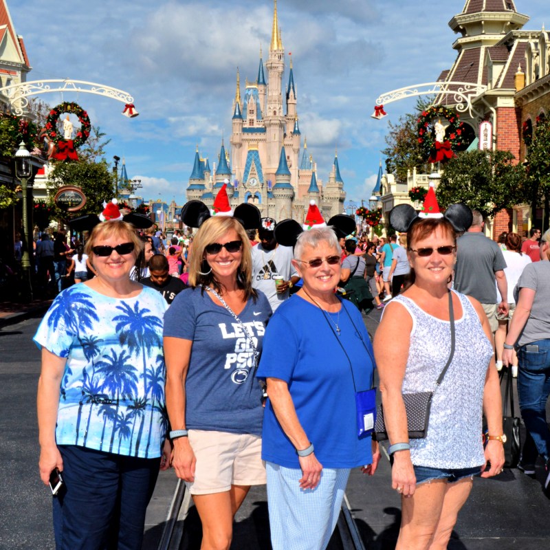 four women wearing mickey ears with santa hat pose in front of cinderella's castle at disney world