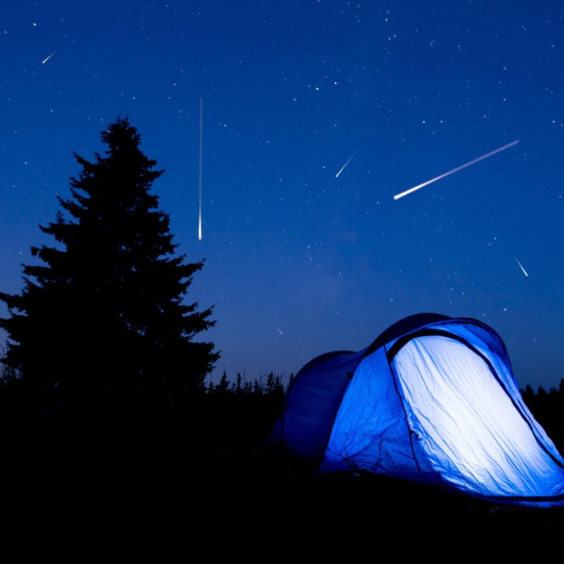 Meteors in the night sky behind an illuminated blue tent