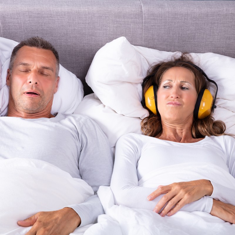 Mature Woman Covering Her Ears With Headphone While Man Snoring In Bed