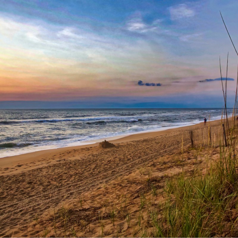 Colorful sunrise on a North Carolina beach, waves breaking on a sandy shore framed by dune grass. Duck, NC