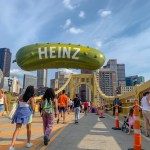 People walking on Andy Warhol Bridge with giant Heinz pickle balloon in the background