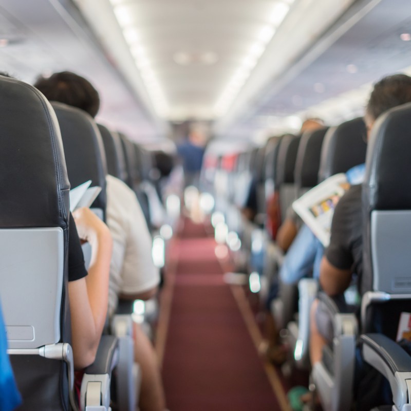 passenger seat, Interior of airplane with passengers sitting on seats and stewardess walking the aisle in background.