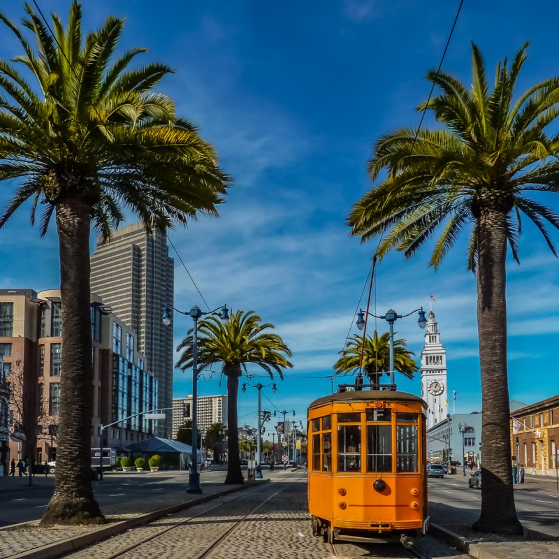 An old orange San Francisco cable car on the Embrcadero with the Ferry Building and palm trees in the background.