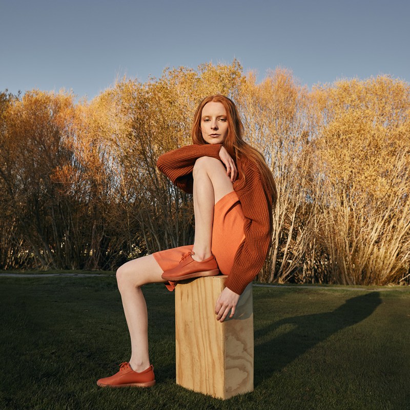 woman with red hair wearing orange sitting on stool in front of fall trees
