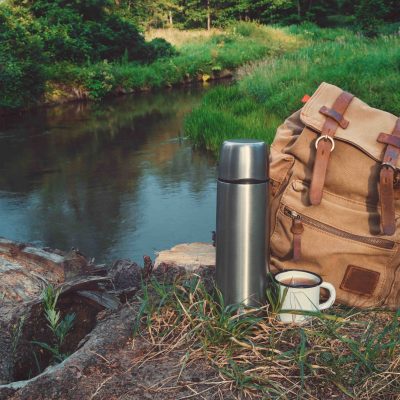 Travel backpack sitting near river next to thermos and coffee mug