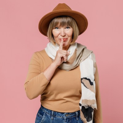 Traveler tourist secret mature elderly senior lady woman 55 years old wears brown shirt hat scarf say hush be quiet with finger on lips isolated on plain pastel light pink background studio portrait